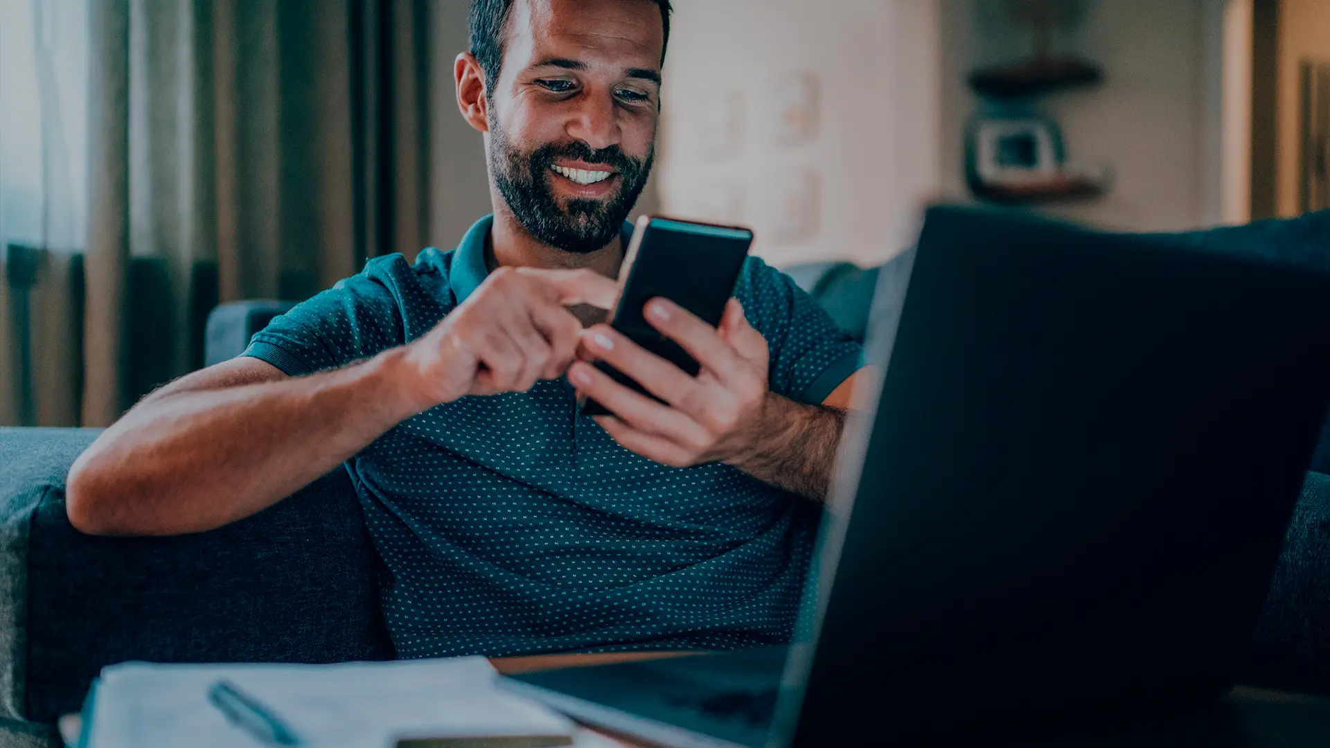 Homem sorrindo enquanto usa o celular em casa, com o notebook ao lado